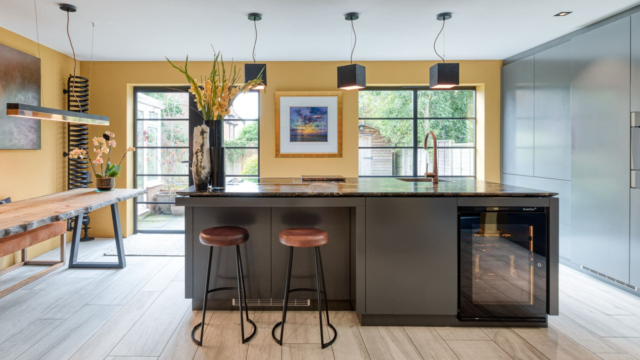 Small glazed service wine cooler, amber lighting, set in a central island of a fitted kitchen in wood and gray colours.