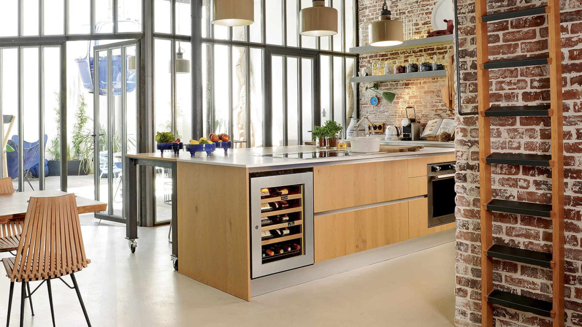 Small size glass door wine cooler with stainless steel frame in an open plan kitchen with industrial design bay window, brick wall and light wood furniture.